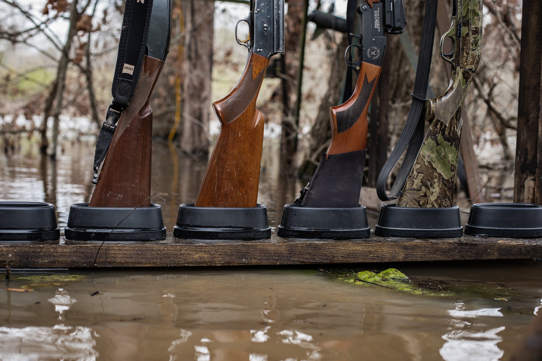 Shotguns standing upright in a row of CoJo Gun Assperatus stock holders in a flooded-timber duck blind